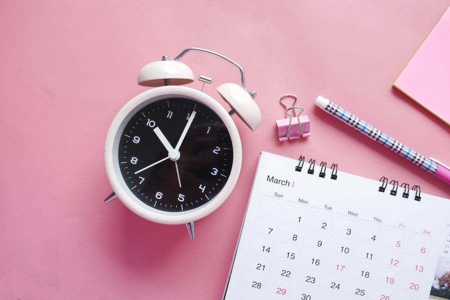 Flat lay of a pink-themed workspace featuring an alarm clock, calendar, pen, and paper clip on a pastel surface.